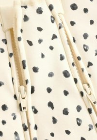 Sweat à capuche en tissu tricoté avec un fond crème orné de taches noires et grises, deux fermetures éclair latérales, et une texture lisse.