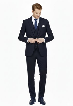 Hombre abrochándose un traje de tres piezas azul marino oscuro con camisa blanca, corbata azul con lunares, pañuelo blanco en el bolsillo y zapatos de vestir negros sobre fondo blanco.