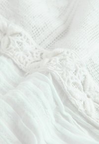Tissu blanc avec un tricot texturé et une finition en dentelle brodée. Comprend des sections douces plissées et une surface lisse et légère.