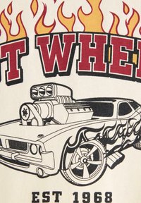 Valkoinen kangas, jossa on graafinen kuva klassisesta autosta, jossa on puhallin, ympäröitynä punaisista ja oransseista liekkikuoseista. Tekstissä lukee "Hot Wheels" ja "Est 1968."