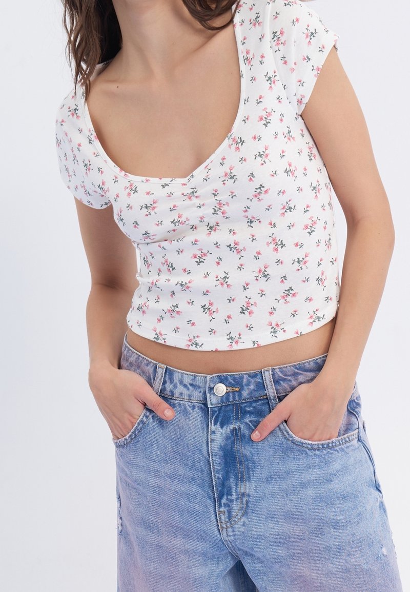 Mujer vestida con un top corto blanco con flores y jeans de tiro alto en azul claro, con las manos en los bolsillos frente a un fondo blanco.