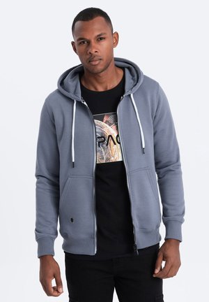 Veste à zip grise avec capuche à cordon, poches avant, manches côtelées, associée à un T-shirt graphique noir avec un design coloré.