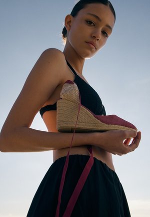 Mladá žena v čiernom topu a sukni drží červenú klinovú espadrilu s viazaním okolo členku na pozadí jasnej oblohy.