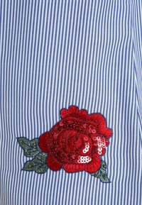 Niebiesko-biała materiał w paski z dużym czerwonym haftem róży wyszywaną cekinami oraz zielonymi liśćmi, z teksturowanym wykończeniem i szczegółowym szyciem.