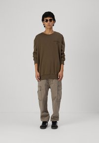 Felpa oversize marrone con logo, abbinata a pantaloni cargo beige con tasche laterali e sneaker nere. Gli accessori includono occhiali da sole.