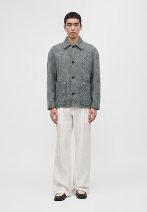 Homme debout portant une veste grise texturée boutonnée, un pantalon blanc ample et des chaussures noires et beiges sur un fond clair uni.