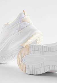Białe plecione sneakersy o teksturowanym wykończeniu, z jednolitą białą podeszwą i miękkim, jasnobeżowym akcentem na pięcie oraz krawędziach podeszwy.