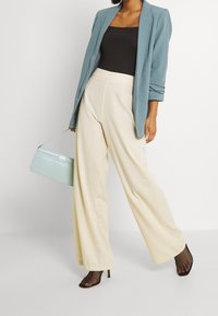 Kobieta w jasnoniebieskiej marynarce, czarnej bluzce, kremowych szerokich nogawkach, czarnych rajstopach w siatkę oraz trzymająca małą jasnoniebieską torebkę.