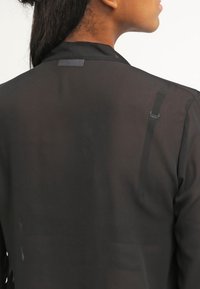 Blouse noire en tissu transparent avec une texture douce, dotée d'un col et d'une discrète fermeture réglable à l'arrière. Design minimaliste.