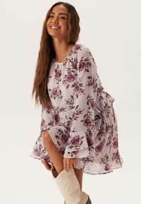 Blommig miniklänning i lätt chiffong, med en lila bas och lila rosiga mönster, utsvängda ärmar och volang nederkant. Kombineras med vita stövlar.