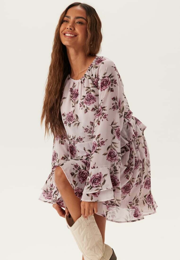 Blommig miniklänning i lätt chiffong, med en lila bas och lila rosiga mönster, utsvängda ärmar och volang nederkant. Kombineras med vita stövlar.