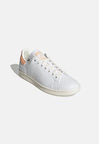 White leather sneaker with a textured finish, orange accent at the heel, and minimalistic design. Features laces and branded tag on the tongue.