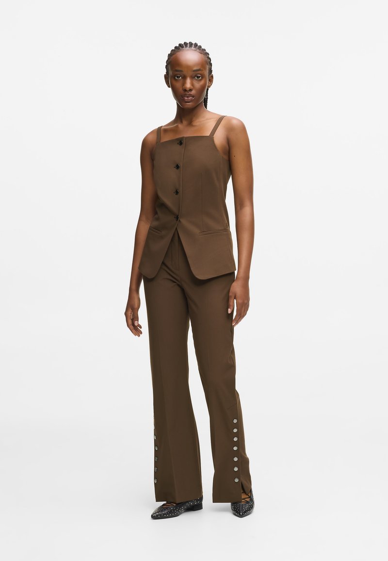 Vest de culoare maro, croit, cu nasturi negri, asociat cu pantaloni asortați care au detalii cu nasturi pe lateral. Material neted și design ajustat.