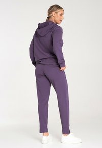 Fioletowa bluza z kapturem i dopasowane dresy z miękkiego materiału, z kapturem, kieszeniami i luźnym krojem. Noszone z białymi trampkami.