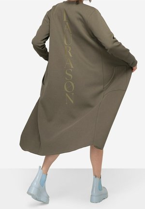 Femme portant un long manteau vert olive avec "LAURASON" imprimé verticalement dans le dos, associée à des bottines bleu clair, en plein mouvement.