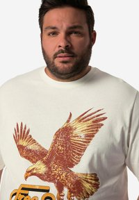 Camiseta de algodón blanca con un gran diseño texturizado de un águila marrón y dorada y el texto "Libre". Cuello redondo, corte relajado.