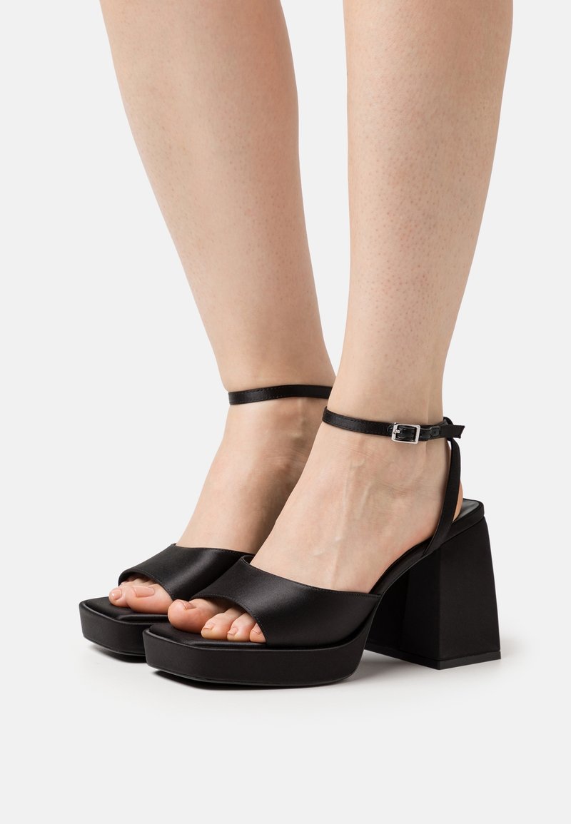 Pieds féminins portant des sandales à talons hauts à plateforme noires avec brides à la cheville et orteils ouverts sur un fond clair uni.