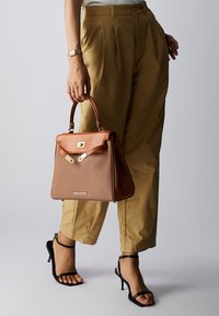 Sac à main marron avec un design structuré, des accents en cuir beige, des détails en métal et une double poignée. Stylisé avec un pantalon beige et des talons noirs.