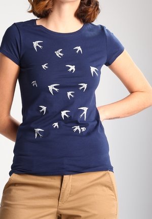 Marineblå tætsiddende T-shirt med hvide svalertryk båret af en person med hænderne bag ryggen, kombineret med beige bukser.