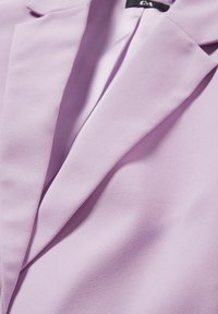 Blazer violet clair avec un col à revers cranté et une texture de tissu lisse, montrant la partie supérieure de la poitrine et la zone du décolleté.