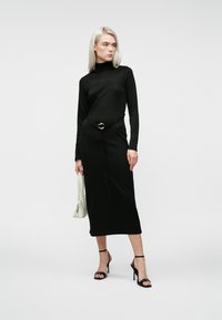 Žena s blond vlasmi, oblečená v čiernych midi šatách s dlhým rukávom a opaskom, drží bielu kabelku a má na sebe čierne lodičky na vysokom opätku.