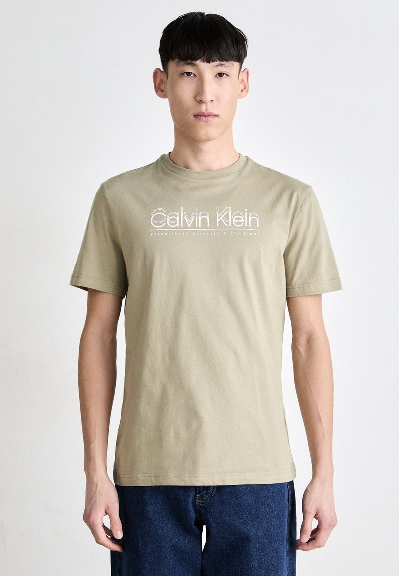 Olivově zelené bavlněné tričko s krátkým rukávem, na kterém je bílý nápis "Calvin Klein" a pod ním "Založeno v roce devatenáct set šedesát osm".
