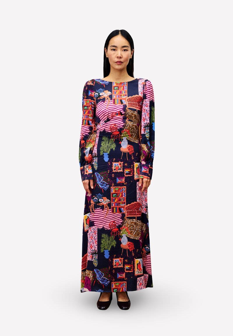 Eine Frau steht in einem langärmeligen Maxikleid mit bunten, abstrakten Mustern, die Pflanzen, Bücherregale, Stühle und gerahmte Bilder zeigen.