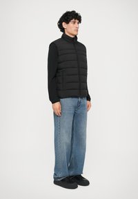 Chaleco de plumas negro con costuras verticales, cuello alto y dos bolsillos laterales. Llevado sobre una camiseta de manga larga negra y combinado con unos pantalones vaqueros de pierna ancha azul.