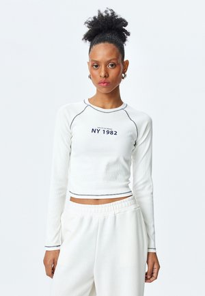 Weißes langärmliges Oberteil mit Rippstruktur und marineblauen Nähten, versehen mit dem Text "NY 1982" und einem cropped Design. Kombiniert mit leichten, lockeren weißen Hosen.