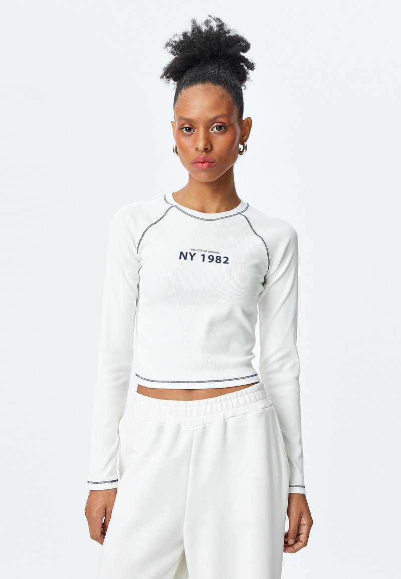 Weißes langärmliges Oberteil mit Rippstruktur und marineblauen Nähten, versehen mit dem Text "NY 1982" und einem cropped Design. Kombiniert mit leichten, lockeren weißen Hosen.