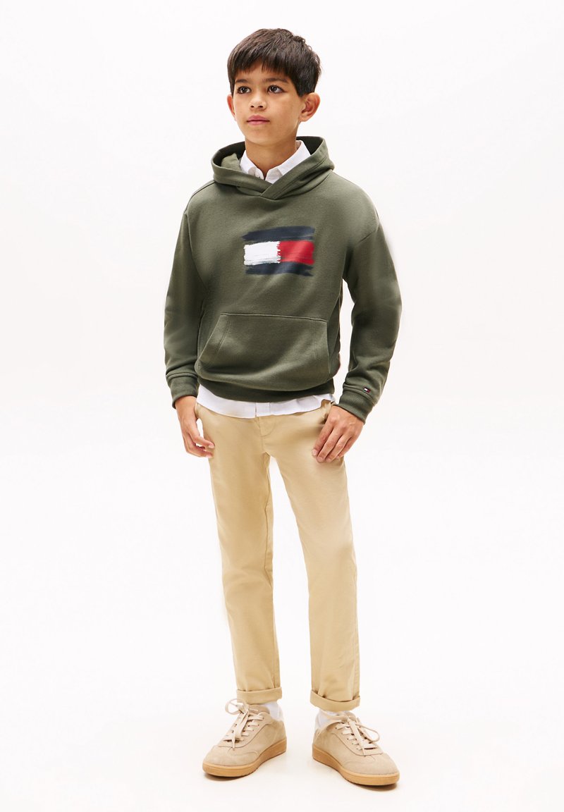 Grøn sweatshirt med et hvidt og rødt abstrakt design på brystet, parret med beige chinos og lysegrå sneakers. Hvid skjorte med krave under.
