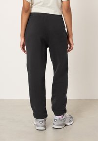 Juodi medvilniniai sportiniai kostiumo kelnės su siaurėjančiomis kojomis, elastiniu juosmeniu ir šoninėmis kišenėmis, dėvimi su pilkais sportiniais batais. Lygus paviršius.