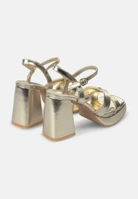 Sandali con tacco a blocco chunky di colore oro metallico, design a cinturino incrociato e fibbia alla caviglia regolabile. Texture liscia, finitura lucida.
