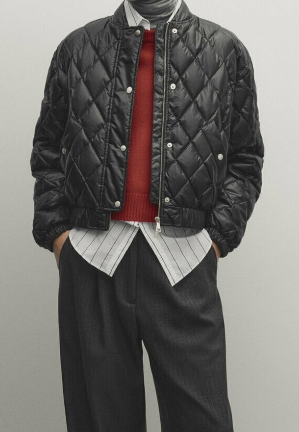 Homme portant une veste bomber matelassée noire sur un pull rouge et une chemise à rayures blanches, associée à un pantalon ample foncé, mains dans les poches.