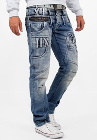 Jean en denim bleu avec plusieurs poches, un délavage usé, des coutures décoratives et une étiquette de marque sur la ceinture; associé à des baskets blanches.