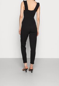 Mono negro con tirantes rizados ajustables, cuerpo ceñido y piernas entalladas. Cierre con cremallera en la parte posterior. Se lleva con sandalias negras de tacón alto.