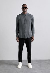Svart-hvitt mønstrete skjorte med lange ermer, kombinert med svarte bukser og hvite joggesko. Modellen har på seg mørke solbriller.