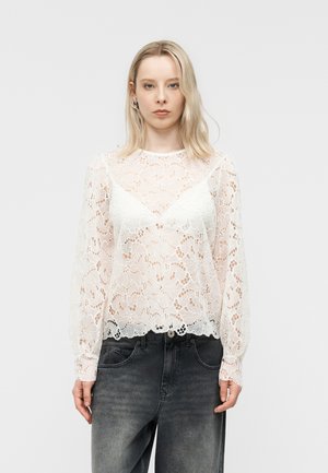 Junge Frau mit glattem blondem Haar, die eine weiße Spitzenbluse mit langen Ärmeln und ausgewaschene schwarze Jeans trägt, schaut nach vorne mit neutralem Gesichtsausdruck.