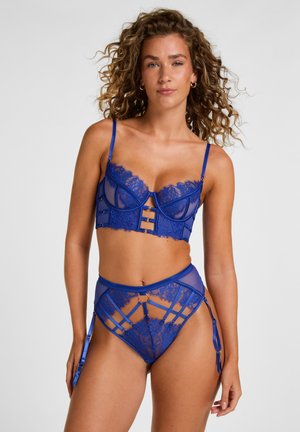 Frau mit lockigem Haar, die ein blaues Spitzen-Dessous-Set mit Strapshaltern trägt, steht vor einem schlichten hellen Hintergrund.