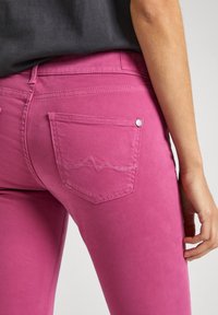 Sittande rosa jeans med en slät yta, med en enda bakficka med en broderad vågmönster och en metallknapp som accent.