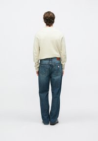 Kremowy sweter na niebieskich jeansach. Jeansy mają brązową skórzaną łatę i dwie tylne kieszenie. Prosty krój z lekko wyblakłym praniem.