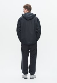 Jachetă impermeabilă neagră cu glugă, asortată cu pantaloni de training negri cu dungi laterale albe. Textură moale, design casual sport.