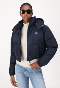Mørkeblå pufferjakke med en cropped design, glidelås lukking og avtagbar hette. Har en logo-patch i rødt, hvitt og blått. Brukt over en beige genser.