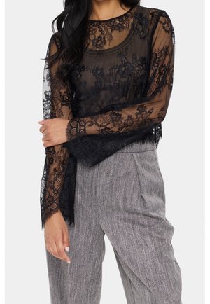 Femme portant un haut transparent en dentelle florale noire à manches longues par-dessus un débardeur noir, associé à un pantalon gris texturé taille haute.