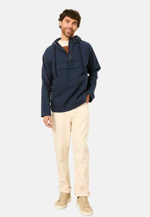 Navy hoodie with a half-zip front, front pocket, and adjustable drawstring. Paired with light beige pants and cream sneakers.