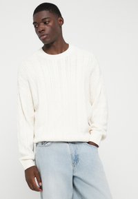 Κρεμ πουλόβερ με πλεξούδα και στρογγυλή λαιμόκοψη, σε συνδυασμό με τζιν ελαφρού πλυσίματος.