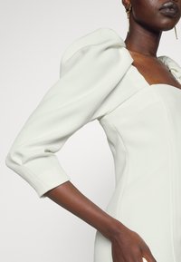 Robe blanche structurée avec des manches bouffantes et un décolleté carré. Elle présente une silhouette ajustée et une texture de tissu lisse.