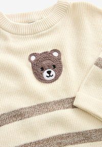 Pull en maille crème avec visage d’ours marron crocheté sur la poitrine et deux bandes horizontales marron sur le devant ainsi qu’une sur le bord de la manche.