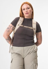 Beige Rucksack mit verstellbaren Trägern, aus strapazierfähigem Material, getragen über einem engen dunklen Oberteil und hellen Cargo-Hosen mit Taschen.