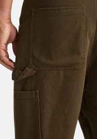 Donkerbruine cargobroek met een gestructureerde stof, zichtbare stiksels, een zijzak en een klein lusje voor gereedschap of accessoires.